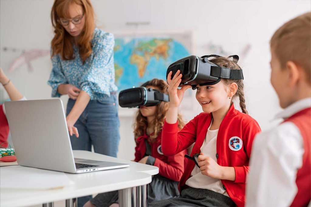 Ein Kind sitzt in der Schule an einem Tisch mit einem aufgeklappten Laptop und hat eine VR-Brille auf dem Kopf sitzen, die sie nach oben schiebt.