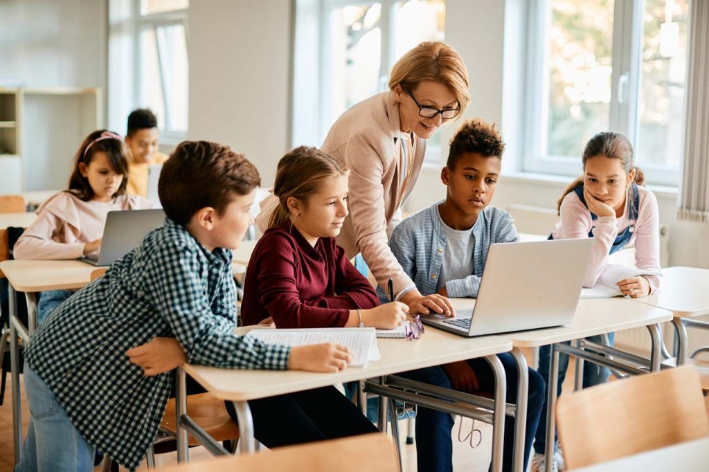 Mehrere Schulkinder schauen auf einen Laptop, während eine Lehrerin hinter ihnen steht und auf dem Laptop etwas zeigt.