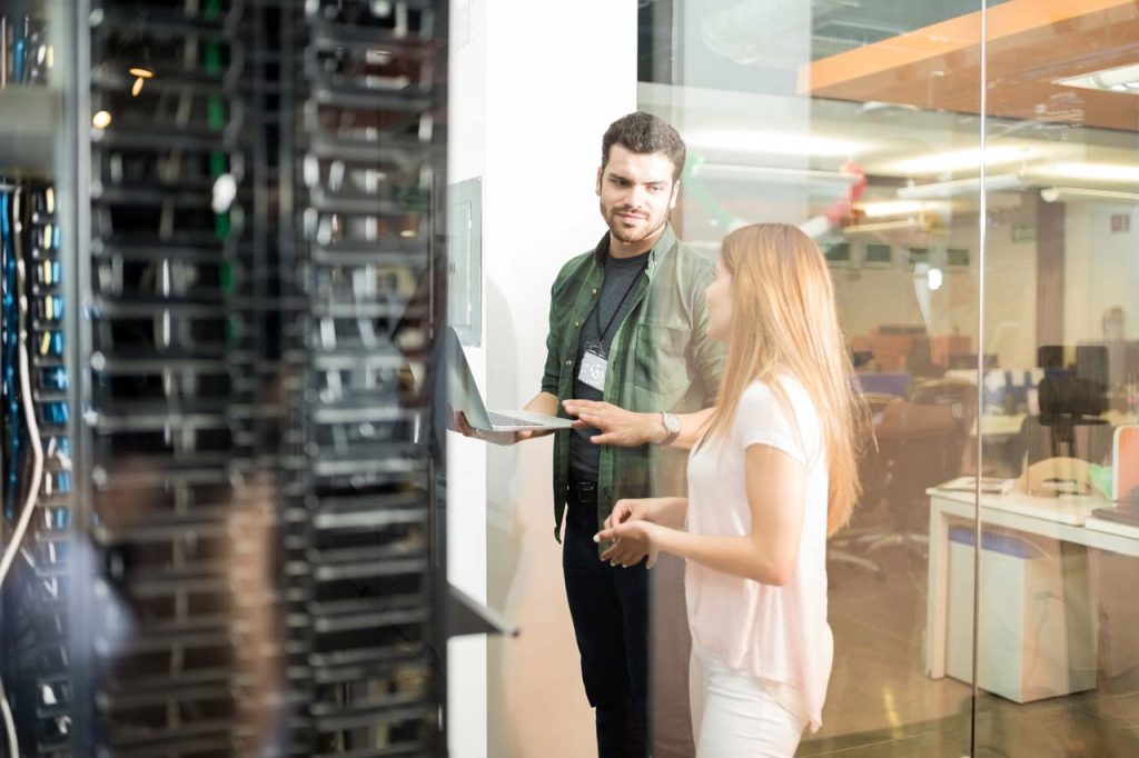 Zwei Personen stehen an einem Server in einem modernen Büro. Ein Mann hält einen Laptop in der Hand während er mit einer Frau neben ihm spricht.